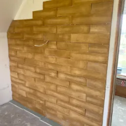 Montaż paneli ściennych imitujących drewno na poddaszu, widoczne niedokończone połączenie z sufitem i wystający przewód elektryczny.