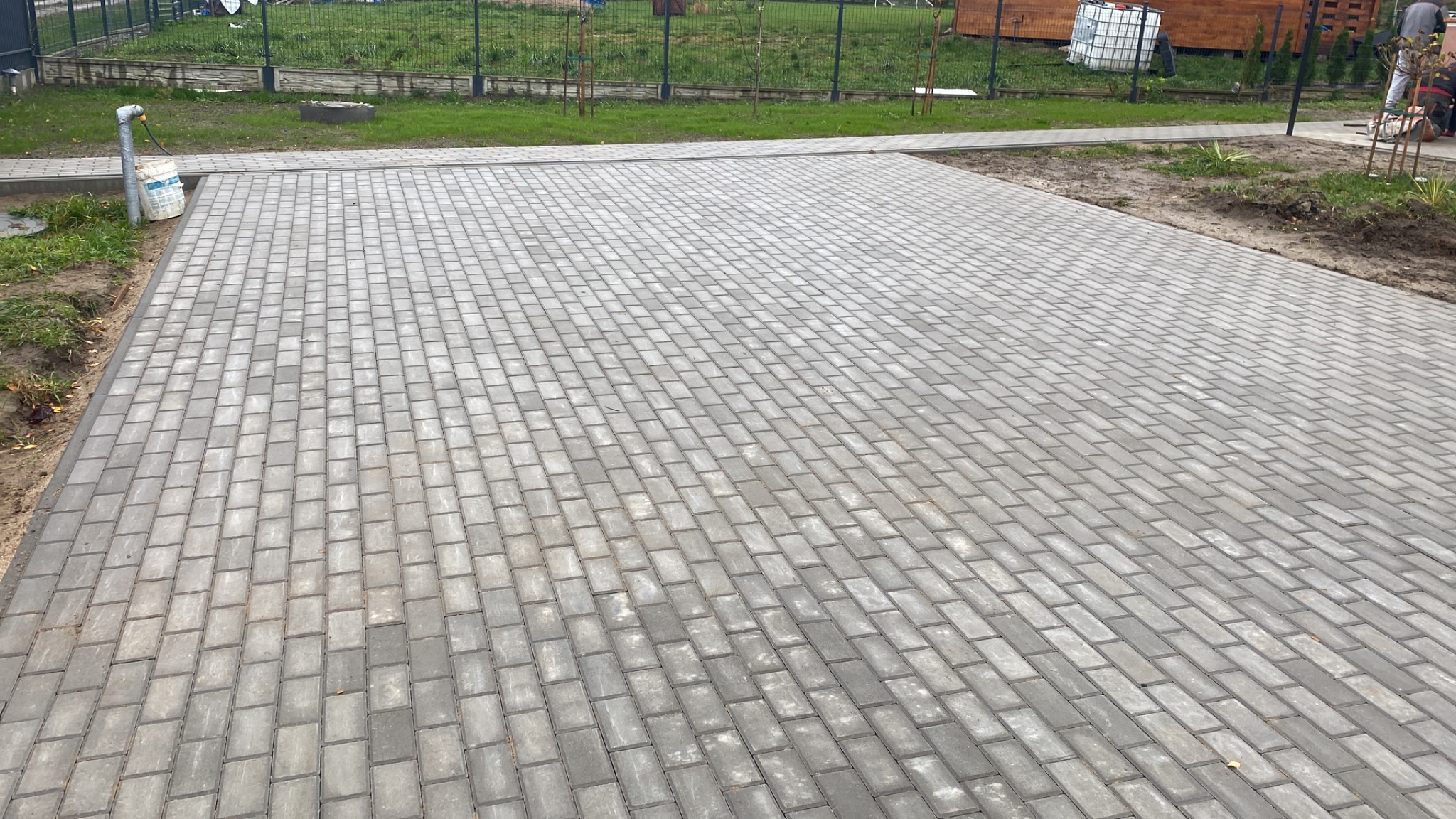 Ułożona szara kostka brukowa na posesji, widoczny fragment trawnika i ogrodzenia w tle.
