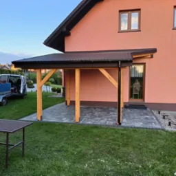 Zadaszenie tarasu przy domu jednorodzinnym z drewnianymi słupami i metalowym dachem, widoczny stół ogrodowy i przyczepka samochodowa.