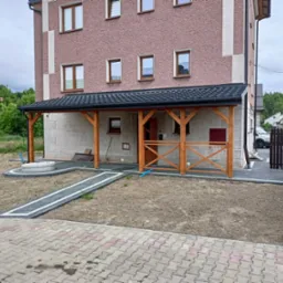 Zadaszony ganek z drewnianą konstrukcją wspartą na słupach, przylegający do dwupiętrowego domu z elewacją w odcieniach różu i szarości, widok z poziomu kostki brukowej.