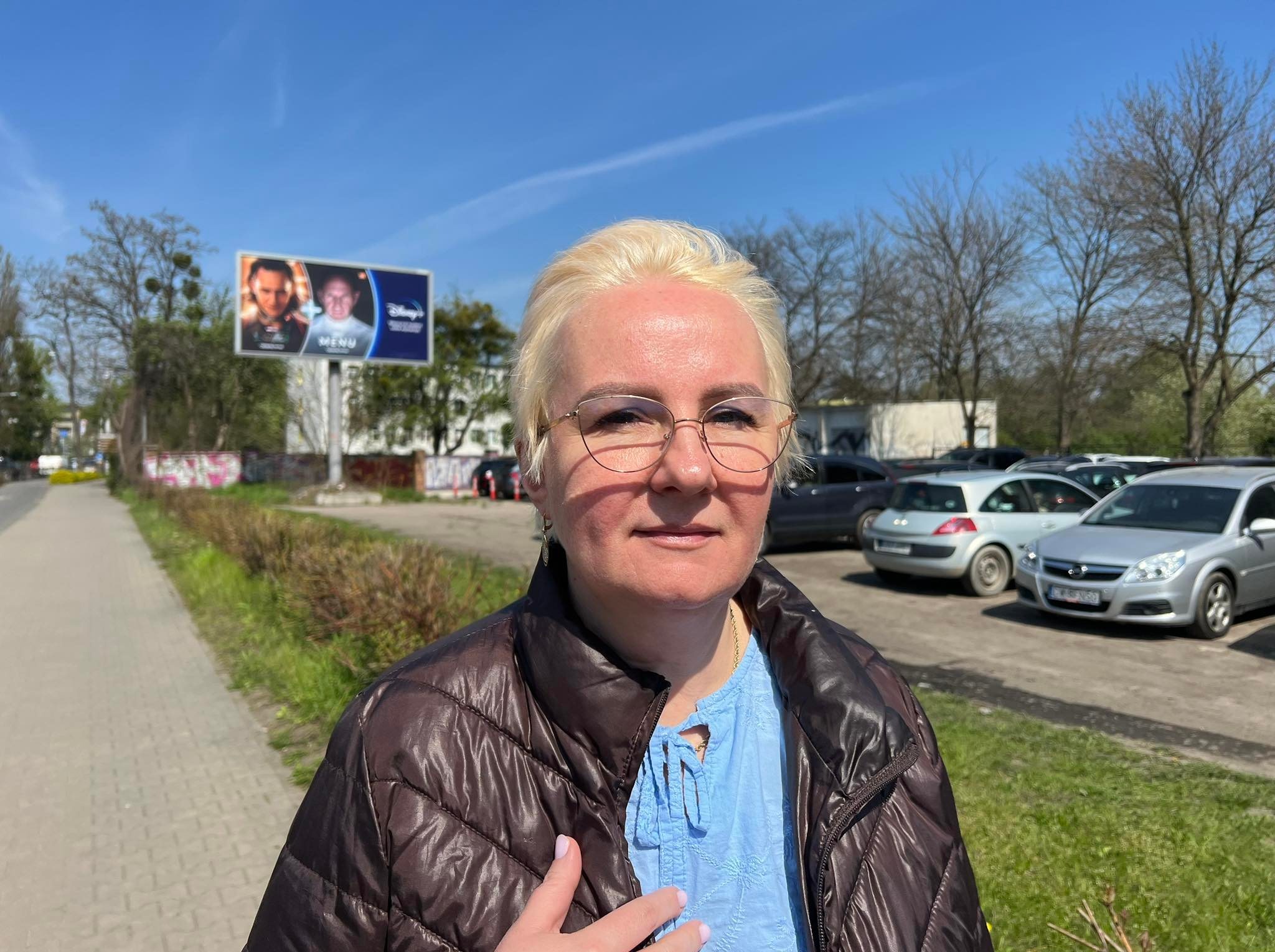 Portret kobiety w okularach i brązowej kurtce na tle parkingu i billboardu Disney Plus.