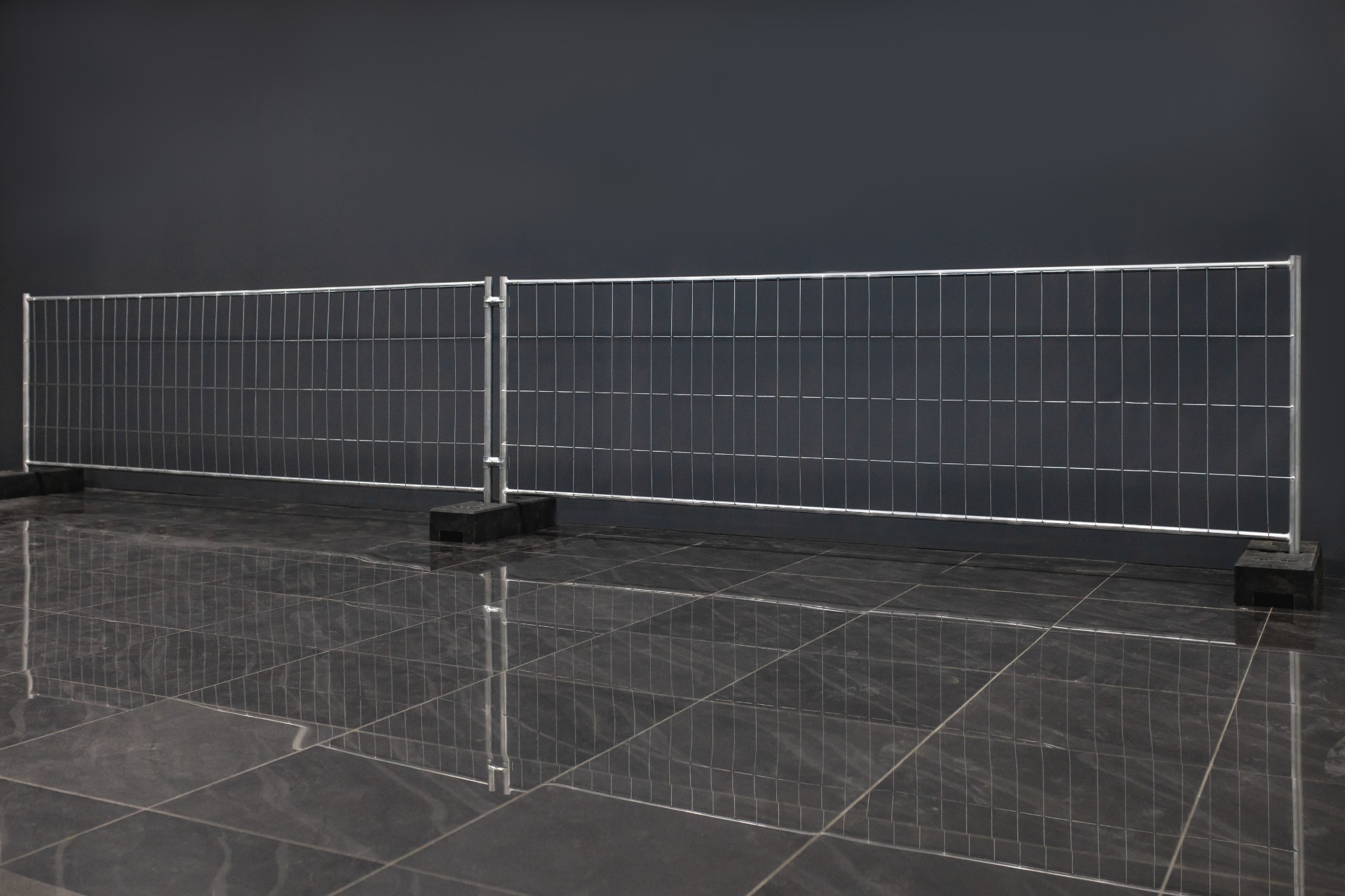 Tymczasowe ogrodzenie panelowe z siatki z metalową ramą i czarnymi podstawami na błyszczącej, ciemnej podłodze, ustawione przed ciemnoszarym tłem.