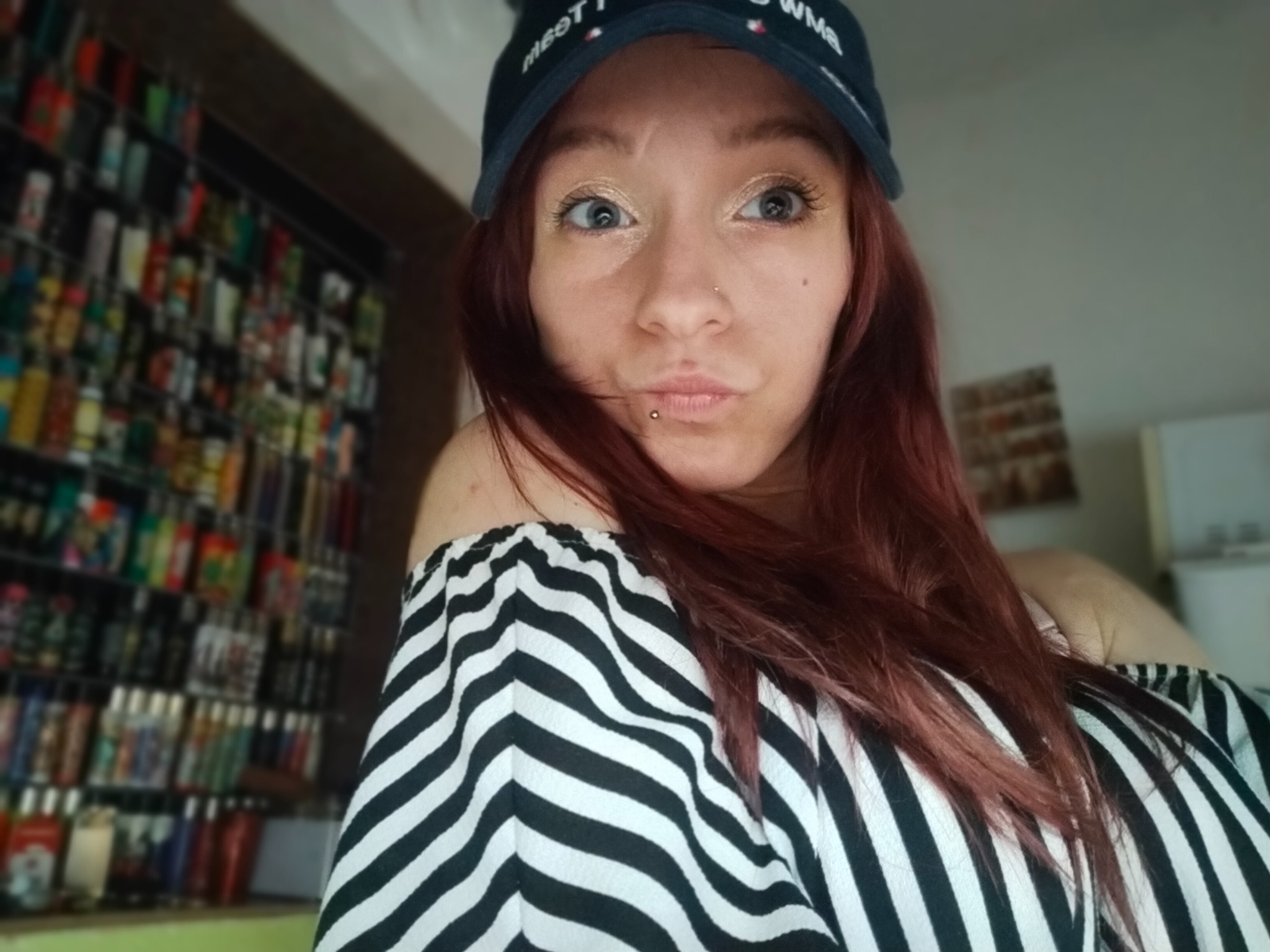 Autoportret kobiety z kolczykiem w wardze i czapce z daszkiem, na tle regału z dużą ilością butelek z płynami.