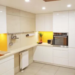 Nowoczesna kuchnia z białymi, lakierowanymi szafkami, żółtym panelem nad blatem, stalowym okapem i relingiem z akcesoriami kuchennymi, uzupełniona grzejnikiem pod blatem.