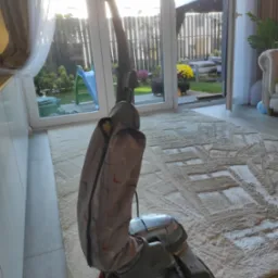 Szaro-beżowy odkurzacz pionowy na tle jasnego dywanu z geometrycznym wzorem, widok z niskiego kąta, z rozsuwanym zamkiem błyskawicznym na obudowie