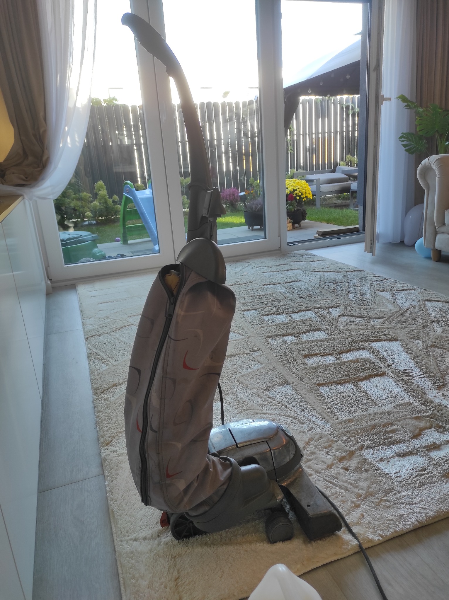 Szaro-beżowy odkurzacz pionowy na tle jasnego dywanu z geometrycznym wzorem, widok z niskiego kąta, z rozsuwanym zamkiem błyskawicznym na obudowie