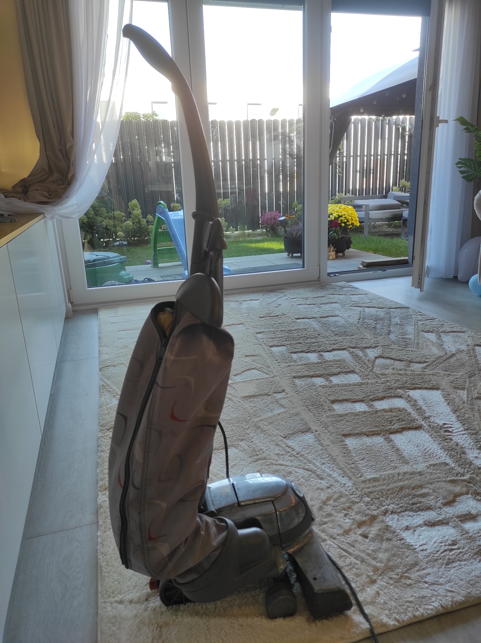 Odkurzacz piorący na beżowym dywanie z geometrycznym wzorem, widok na ogród z altaną przez otwarte drzwi tarasowe.