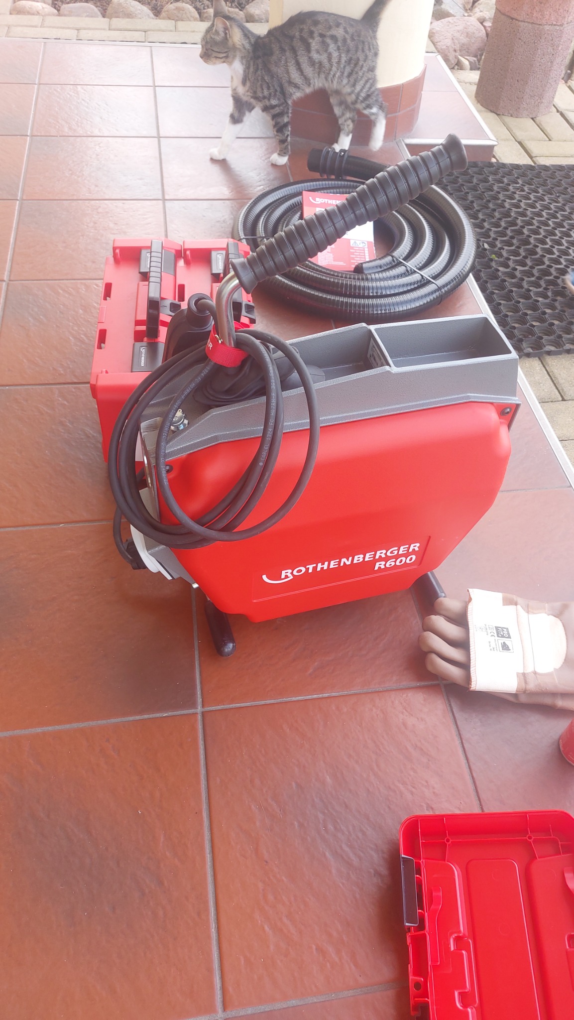 Pomarańczowa maszyna Rothenberger R600 do czyszczenia rur z akcesoriami na brązowych płytkach, z kotem w tle.