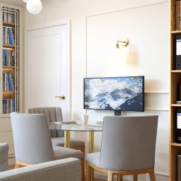 MM-Studio Projektowanie Wnętrz Marcin Martela - Nowoczesny pokój z szarą tapicerką krzeseł przy szklanym stole, na ścianie telewizor z górskim krajobrazem i lampa, obok regał z segregatorami i książkami.