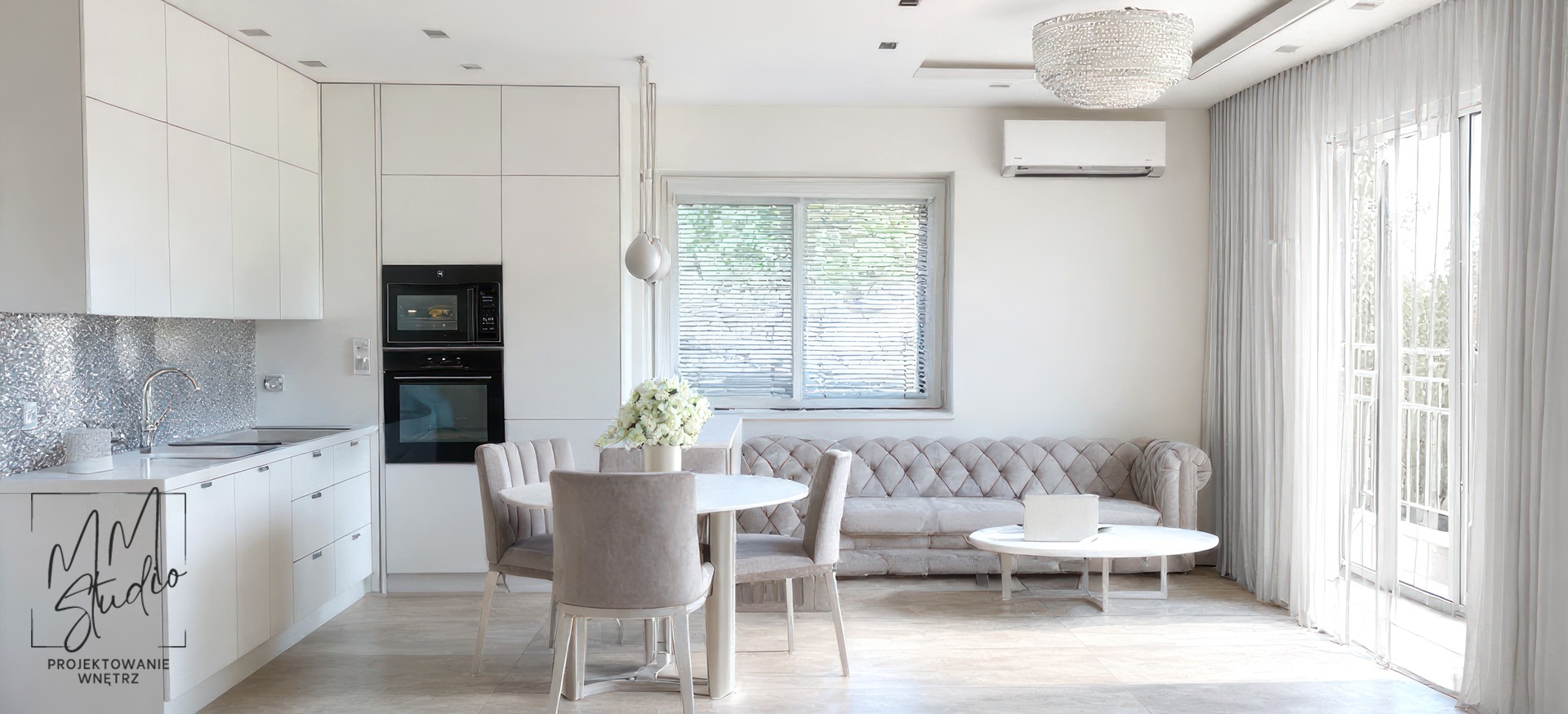 Jasne, nowoczesne wnętrze z aneksem kuchennym, okrągłym stołem i sofą w stylu Chesterfield. Błyszcząca mozaika nad blatem. Lampa kryształowa.