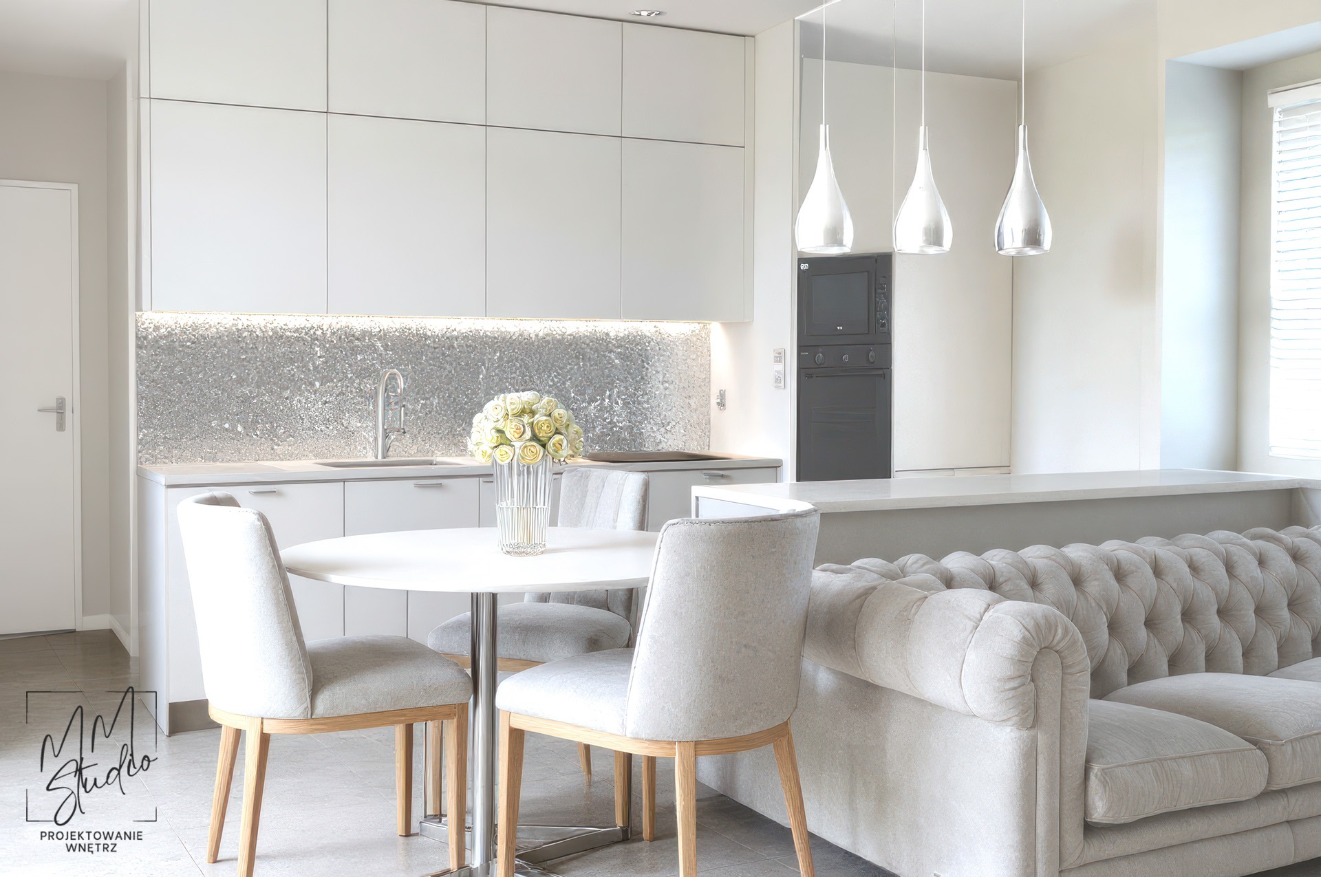 Jasna kuchnia z srebrną mozaiką nad blatem, okrągły stół z kwiatami i fragment sofy w stylu Chesterfield. Nowoczesne, wiszące lampy.