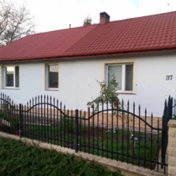 Dom jednorodzinny z ocieploną i pomalowaną na biało elewacją, czerwonym dachem z dachówki, czarnym metalowym ogrodzeniem i numerem 37 na ścianie.