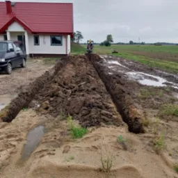 Mini koparka wykonuje wykop pod instalacje na błotnistym terenie obok domu z czerwoną dachówką.