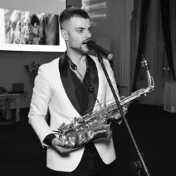 Czarno-białe zdjęcie saksofonisty w białym smokingu śpiewającego do mikrofonu na tle sali bankietowej z wyświetlanym obrazem.