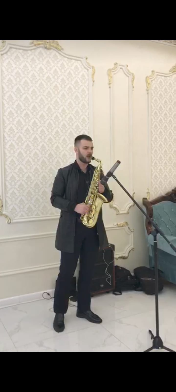 Saksofonista w czarnym stroju gra na złotym saksofonie przed mikrofonem w pomieszczeniu z dekoracyjną ścianą.