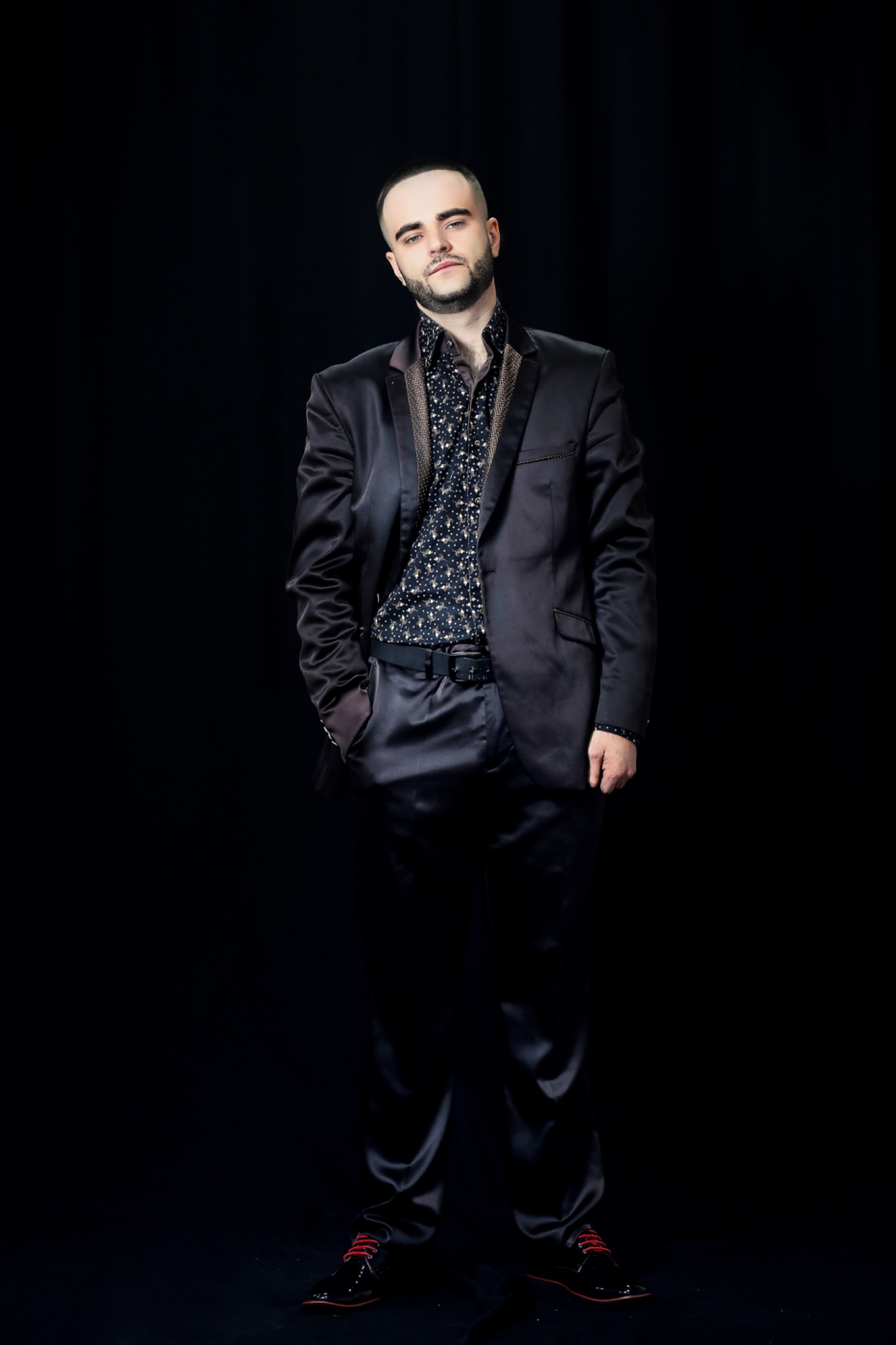 Portret mężczyzny w ciemnym, błyszczącym garniturze i wzorzystej koszuli, pozującego na czarnym tle.
