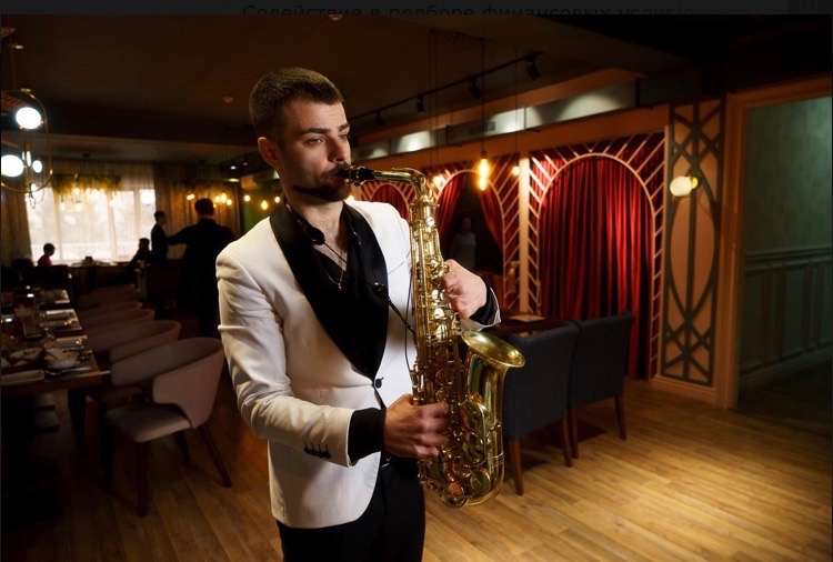 Saksofonista w białej marynarce występuje w restauracji, grając na saksofonie tenorowym, w tle widoczne stoliki i dekoracje wnętrza.