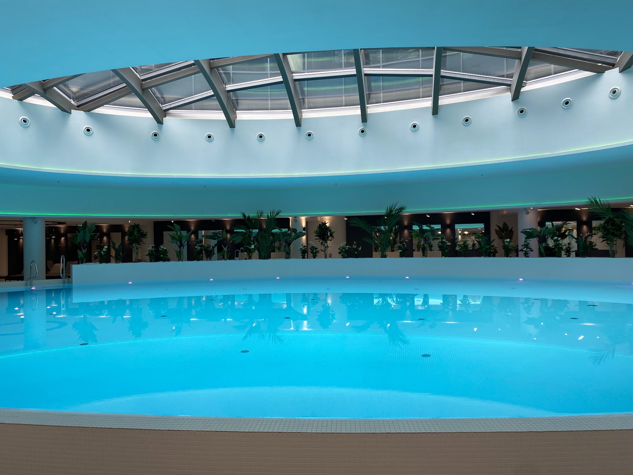 Błękitny basen w nowoczesnym spa z przeszklonym dachem i roślinami w tle.