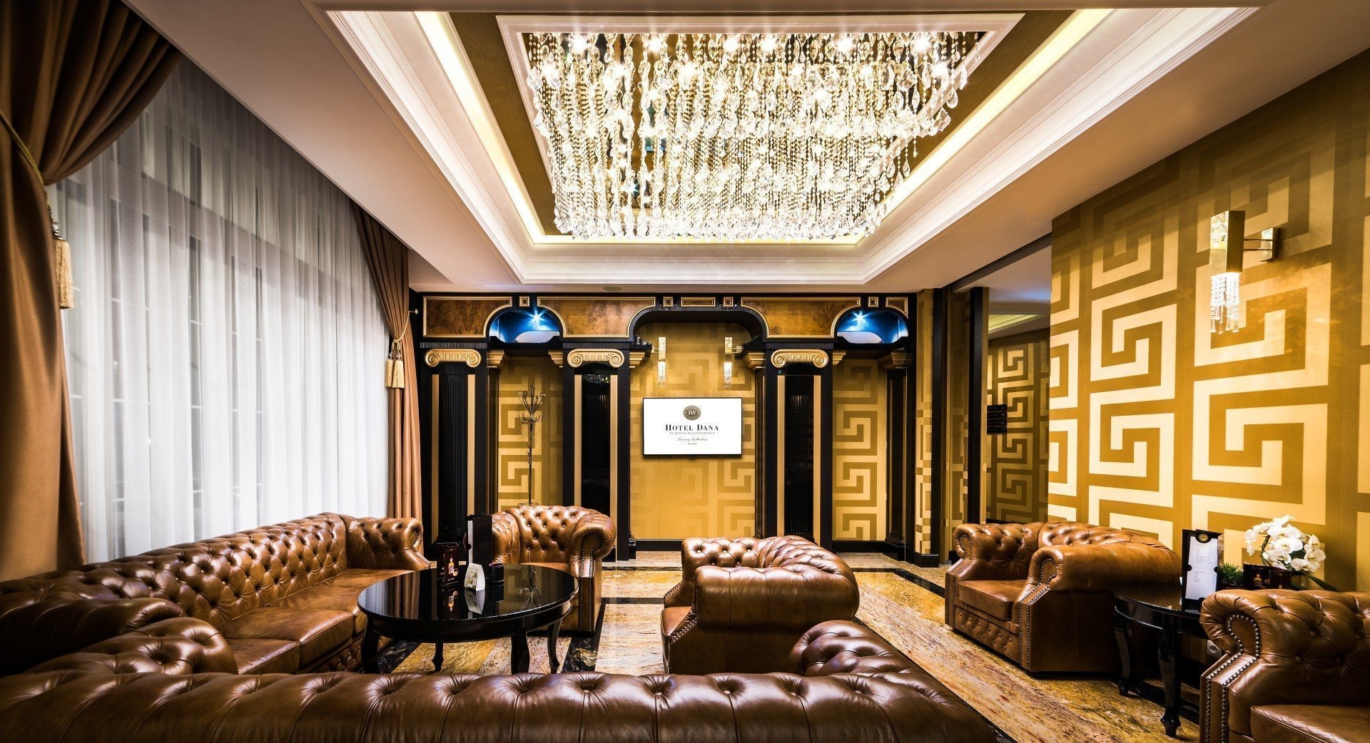 Luksusowe wnętrze hotelowe z brązowymi skórzanymi sofami chesterfield, kryształowym żyrandolem i ścianami w geometryczne wzory, widoczny ekran z logo Hotel Dana.