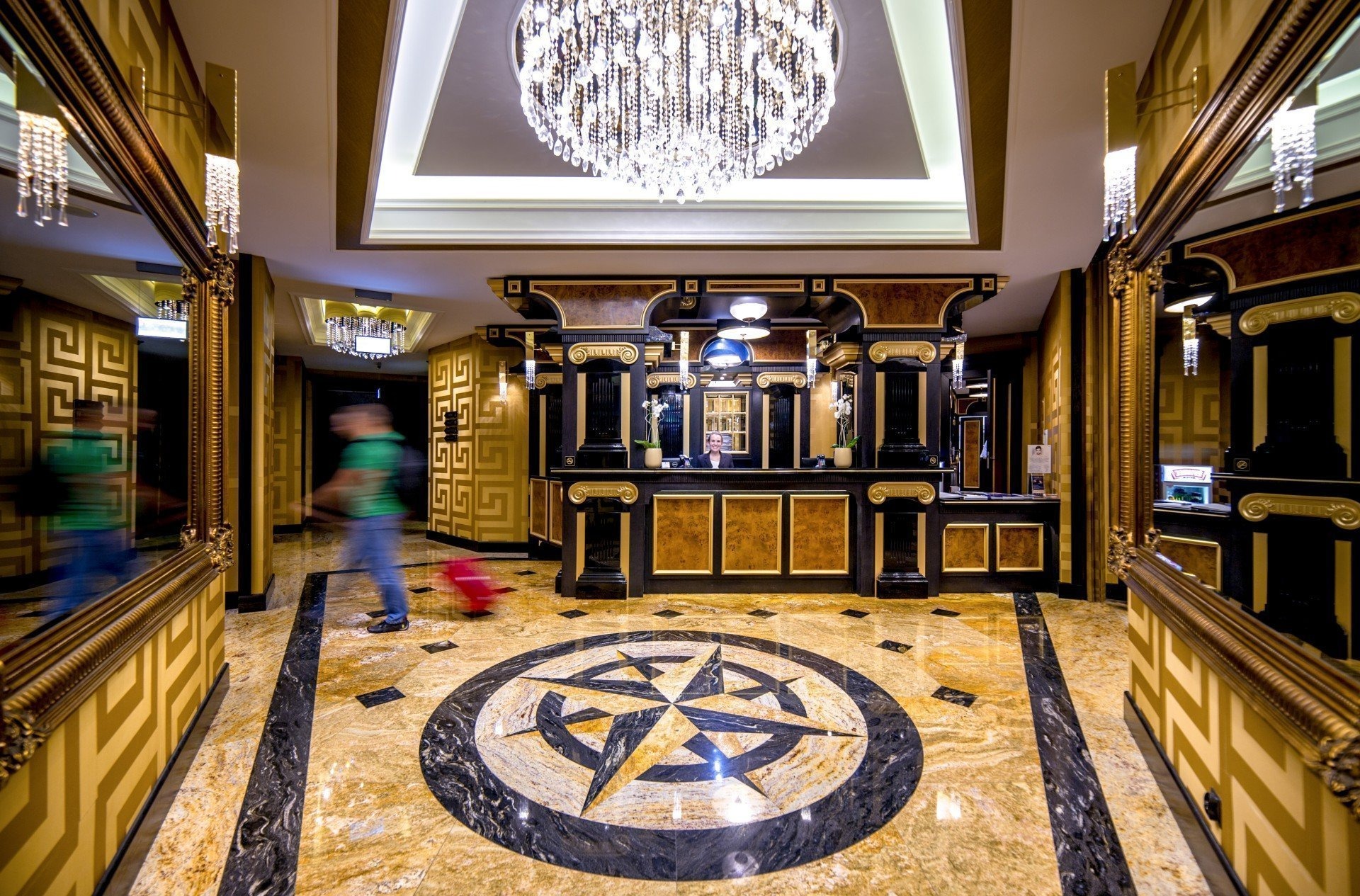 Luksusowe lobby hotelowe z marmurową podłogą w kształcie kompasu, kryształowym żyrandolem i recepcją z ciemnego drewna ze złotymi zdobieniami. Rozmyta postać z czerwoną walizką przechodzi obok...