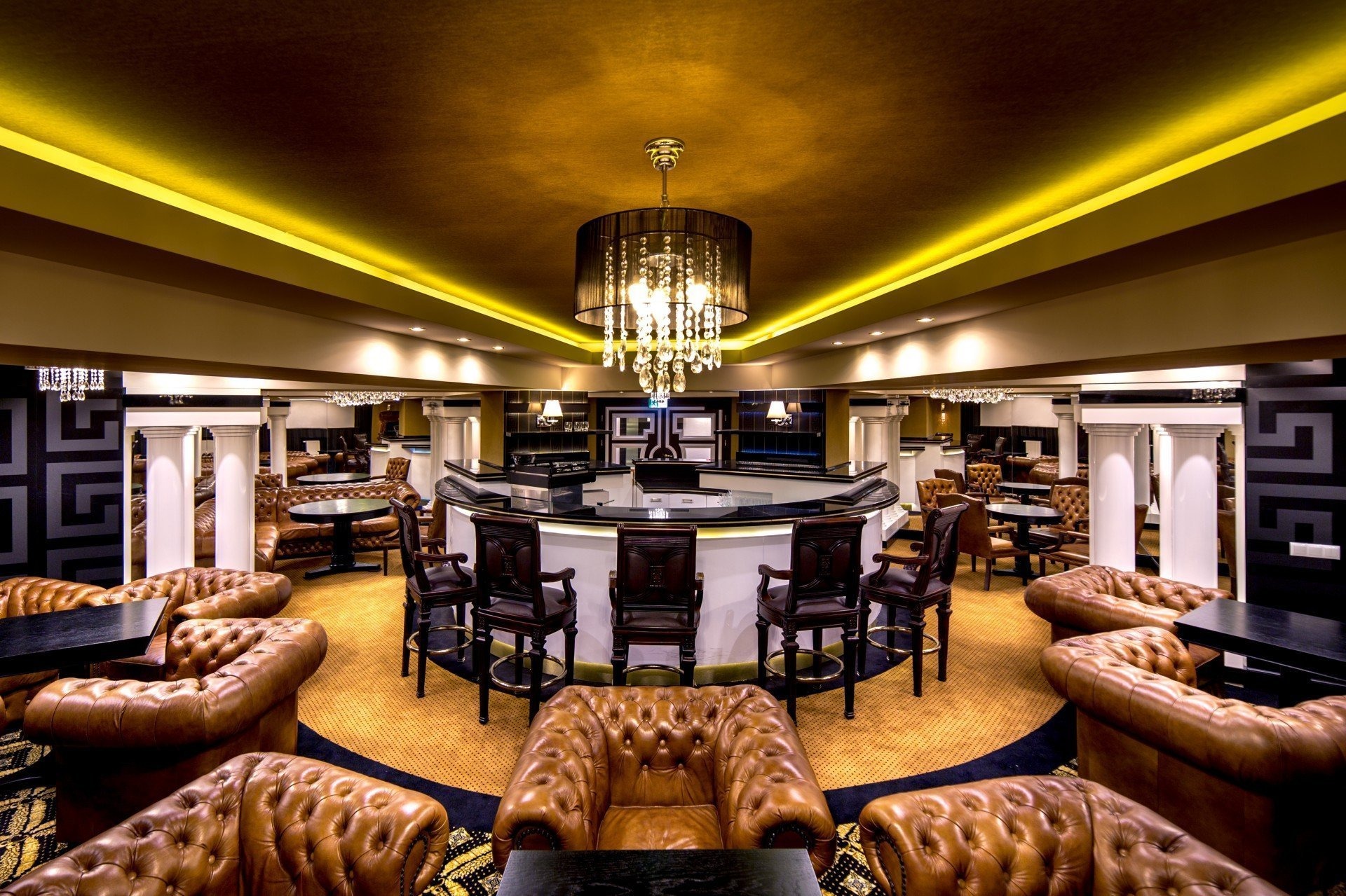 Elegancki bar z okrągłym kontuarem, ciemnymi skórzanymi fotelami chesterfield i kryształowym żyrandolem podświetlonym ciepłym światłem.
