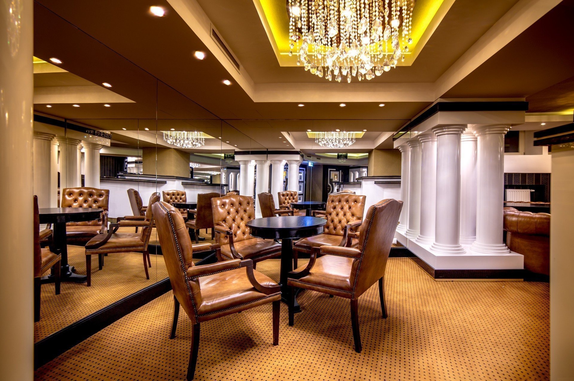 Eleganckie wnętrze sali bankietowej z brązowymi skórzanymi fotelami, okrągłymi czarnymi stolikami, kolumnami i kryształowym żyrandolem.
