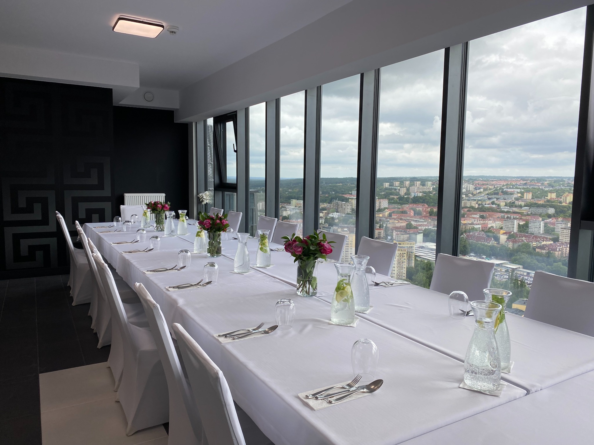 Długi stół z białym obrusem, nakryty dla wielu gości, dekoracje z kwiatów i wody, widok na miasto zza okien panoramicznych.
