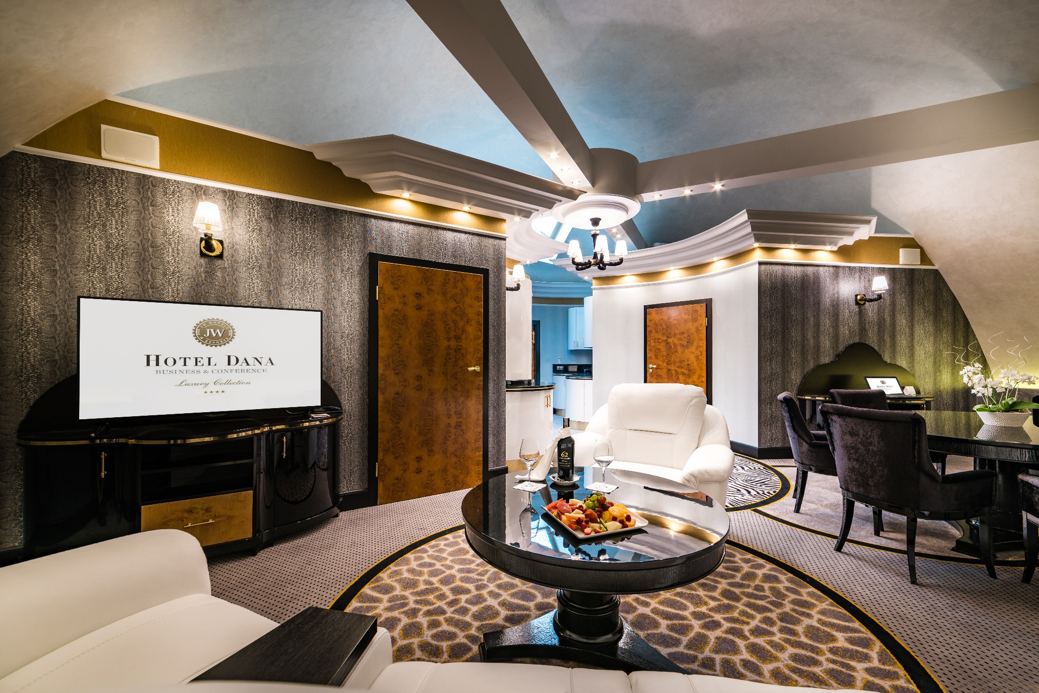 Luksusowy apartament hotelowy z owocami na stoliku kawowym, widok na telewizor z logo hotelu i jadalnię w tle.