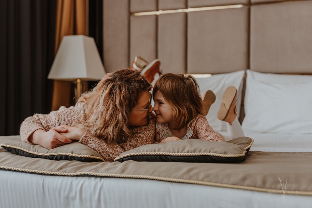 Kobieta w okularach i mała dziewczynka leżą na łóżku w pokoju hotelowym, patrząc na siebie z uśmiechem.