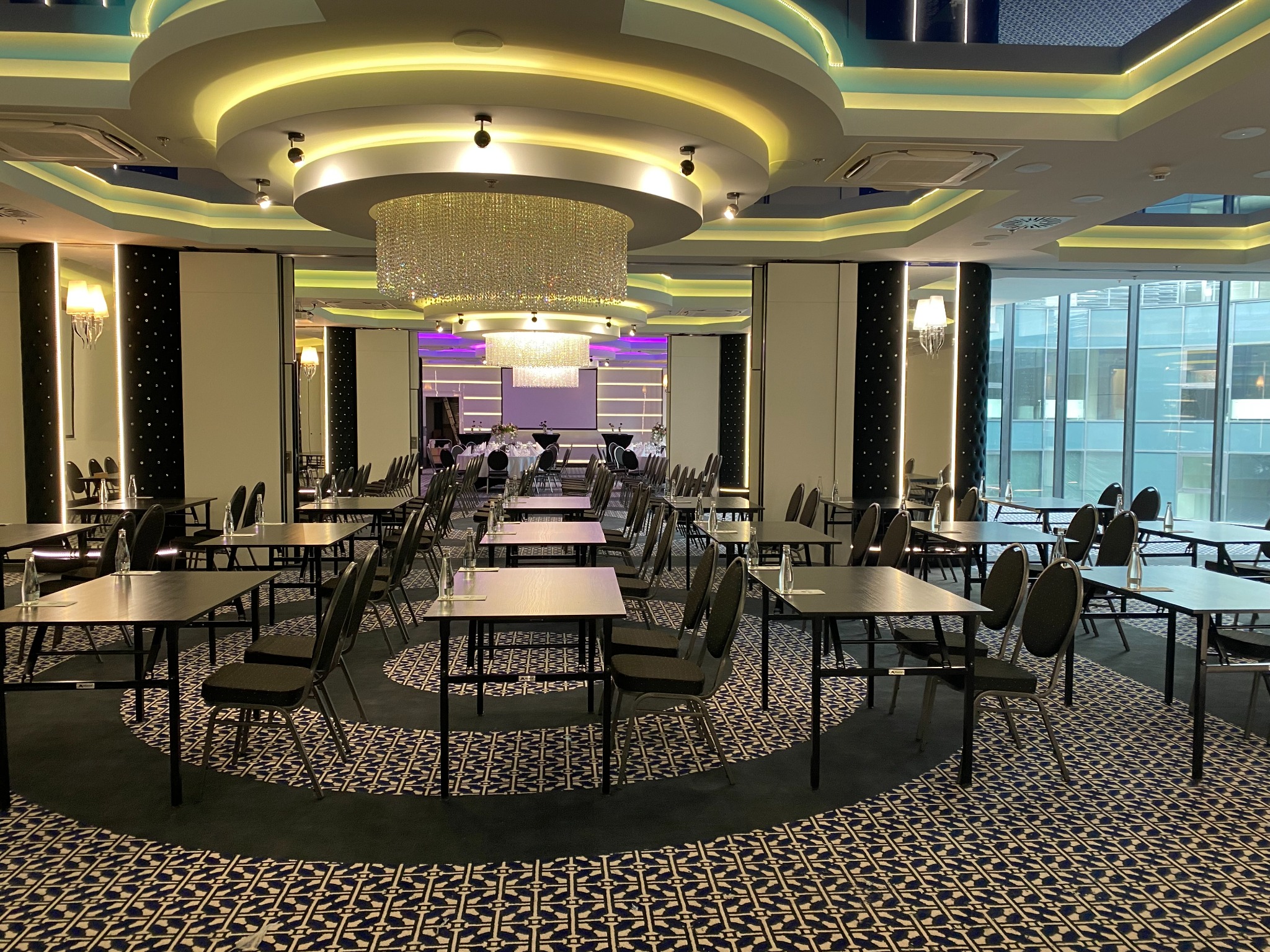 Sala konferencyjna z rzędami stołów i krzeseł, oświetlona ozdobnymi żyrandolami i podświetleniem sufitowym, przygotowana do wydarzenia.