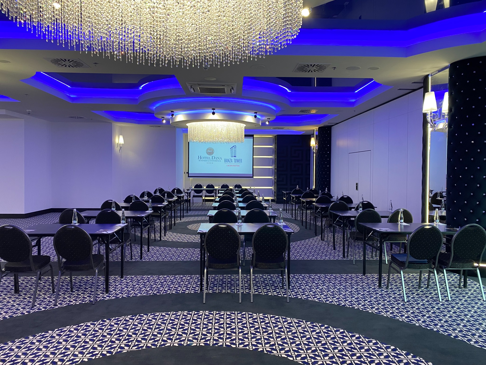 Sala konferencyjna z rzędami czarnych krzeseł i stołów, oświetlona dekoracyjnymi żyrandolami i niebieskim światłem, z butelkami wody na stołach i ekranem z logo hotelu w tle.
