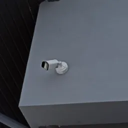 Kamera monitoringu zamontowana na szarej ścianie zewnętrznej budynku, widoczny fragment dachu z blachy trapezowej.