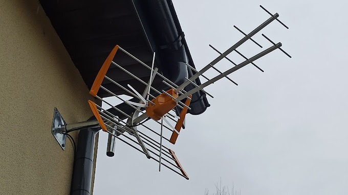 Zewnętrzna antena telewizyjna DVB-T z pomarańczowymi elementami, zamontowana na elewacji domu obok rynny, widok z dołu na tle pochmurnego nieba.