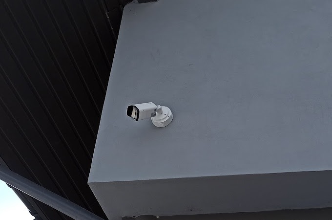 Kamera monitoringu zamontowana na szarej ścianie zewnętrznej budynku, widoczny fragment dachu z blachy trapezowej.