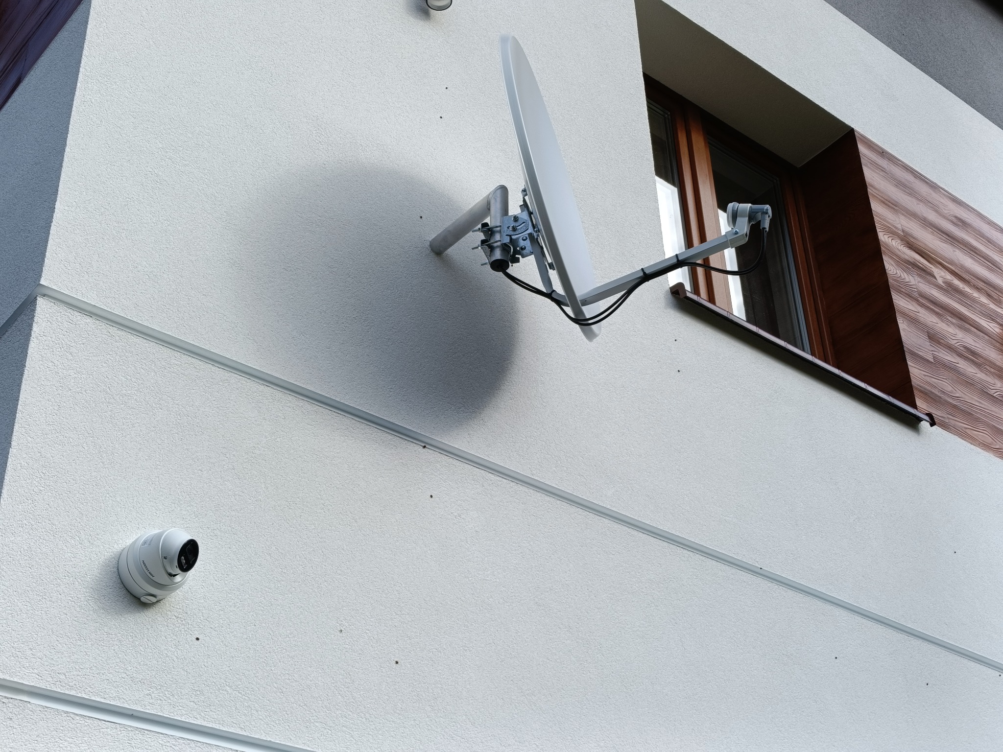 Biała antena satelitarna i kamera monitoringu zamontowane na elewacji budynku z oknem. Widok z dołu, światło dzienne.