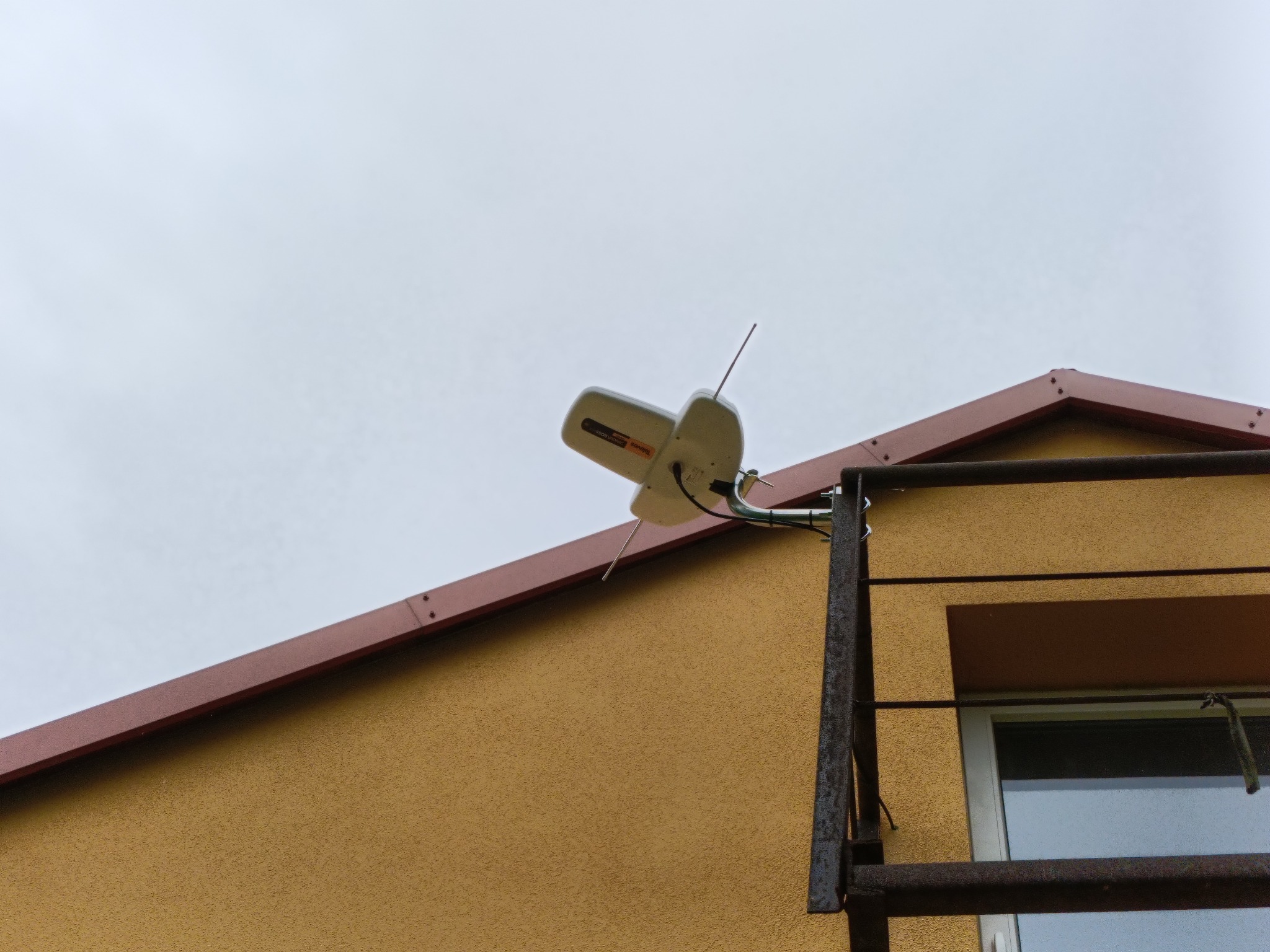 Antena telewizyjna zamontowana na elewacji budynku z drabinką, widok z dołu, pochmurne niebo w tle. Detal montażu anteny.