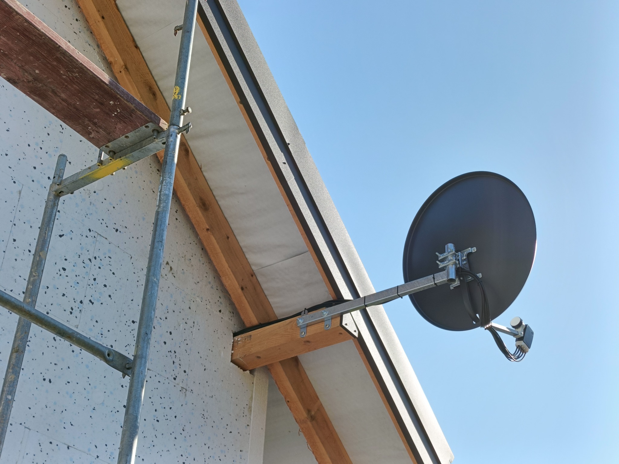 Antena satelitarna zamontowana na elewacji budynku, widoczna z dołu na tle błękitnego nieba. Obok rusztowanie z drewnianą deską.