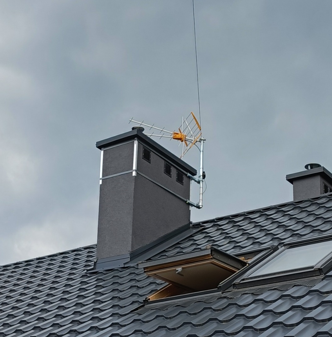Antena telewizyjna zamontowana na kominie domu z grafitową dachówką i oknami dachowymi, na tle pochmurnego nieba. Widok z dołu.