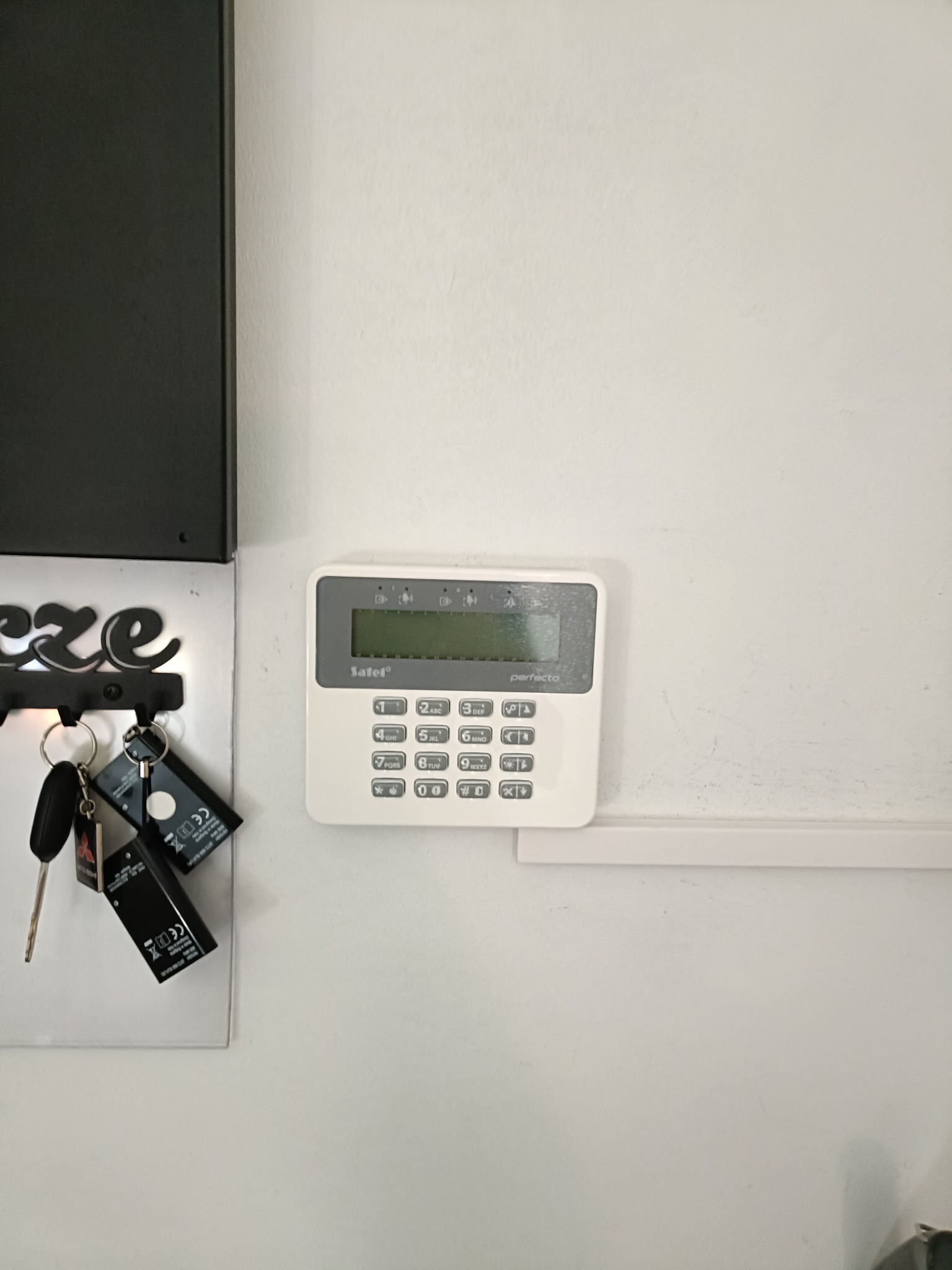 Biały panel sterowania alarmu Safel Perfecto na ścianie, obok wieszak na klucze z kluczami. Widoczne ślady użytkowania na ścianie.