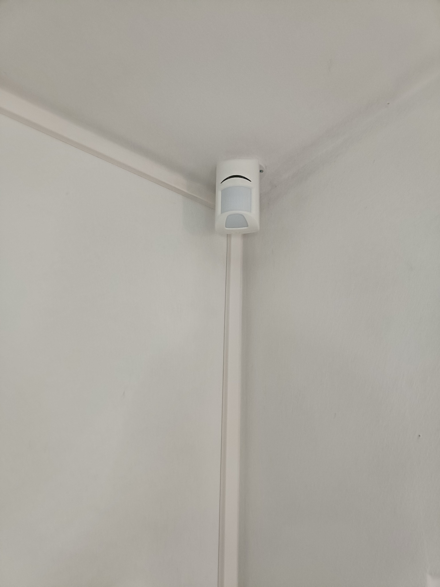 Zamontowany czujnik ruchu w rogu pomieszczenia, przymocowany do listwy przypodłogowej. Białe ściany i sufit. Widok z dołu, blisko rogu.