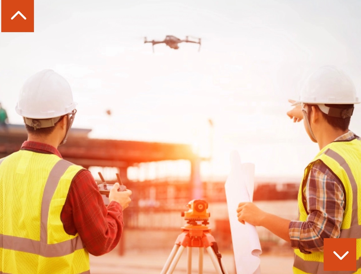 Dwóch geodetów w kamizelkach i kaskach obserwuje drona podczas pomiarów, w tle widoczny niwelator optyczny na statywie, jeden geodeta trzyma kontroler drona, drugi wskazuje palcem na drona...