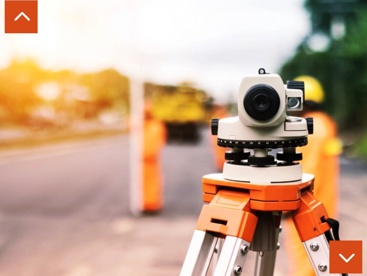 Teodolit ustawiony na trójnogu, widoczni pracownicy w pomarańczowych kamizelkach na drodze w tle, ciepłe światło słoneczne.