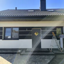 Montaż pompy ciepła Vaillant przy elewacji domu z czarno-białymi poziomymi pasami, widoczna drabina i pracownik przy oknie.