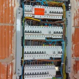 Rozdzielnica elektryczna z zainstalowanymi bezpiecznikami i okablowaniem, osadzona w surowej ścianie z cegły, widoczne elementy montażowe i oznaczenia na urządzeniach.