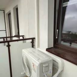 Zewnętrzna jednostka klimatyzacji Rotenso Ukura zamontowana na balkonie z widokiem na zewnątrz, z brązowymi ramami okiennymi i szarą pogodą w tle.