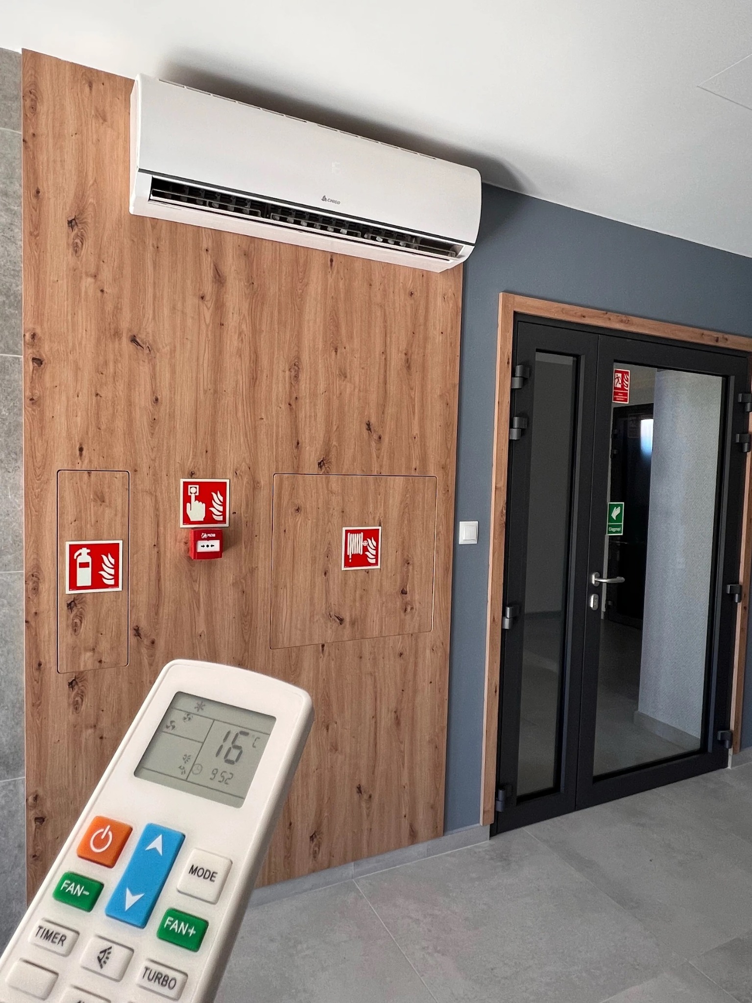 Biała jednostka wewnętrzna klimatyzatora zamontowana na drewnianej ścianie w biurze, sterowana pilotem ustawionym na 16 stopni Celsjusza, widoczne drzwi przeciwpożarowe i oznaczenia bezpieczeństwa.
