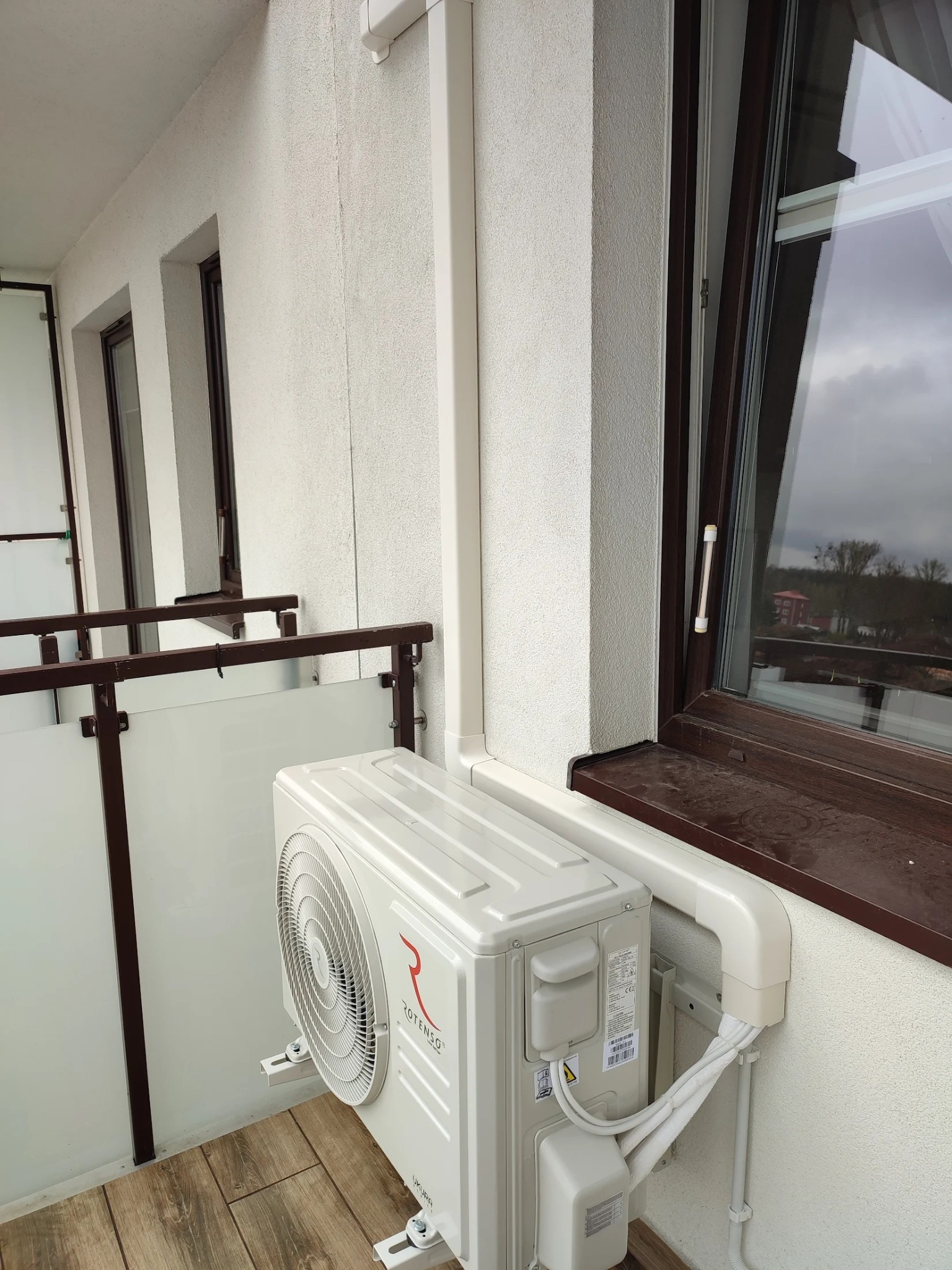 Zewnętrzna jednostka klimatyzacji Rotenso Ukura zamontowana na balkonie z widokiem na zewnątrz, z brązowymi ramami okiennymi i szarą pogodą w tle.