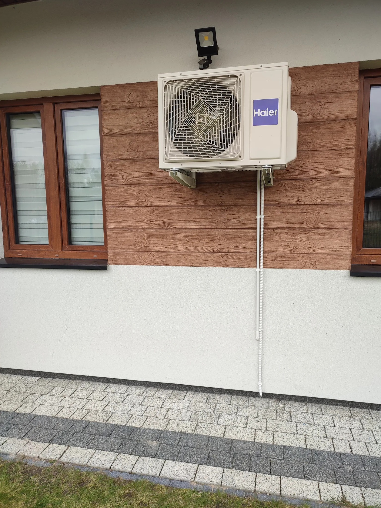 Biała jednostka zewnętrzna klimatyzatora Haier zamontowana na brązowej elewacji imitującej drewno, z widocznymi przewodami w białej listwie, obok okna z brązową ramą i czujnikiem ruchu...