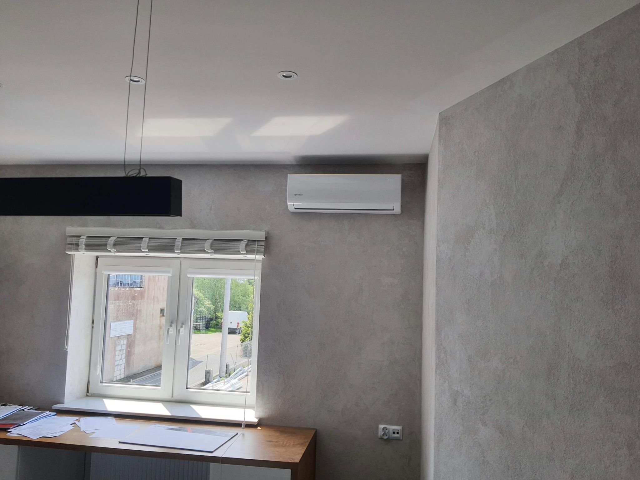 Biała jednostka klimatyzacyjna zamontowana na ścianie nad oknem w pomieszczeniu z szarą, teksturowaną ścianą. Na parapecie leżą dokumenty i biurowe akcesoria.