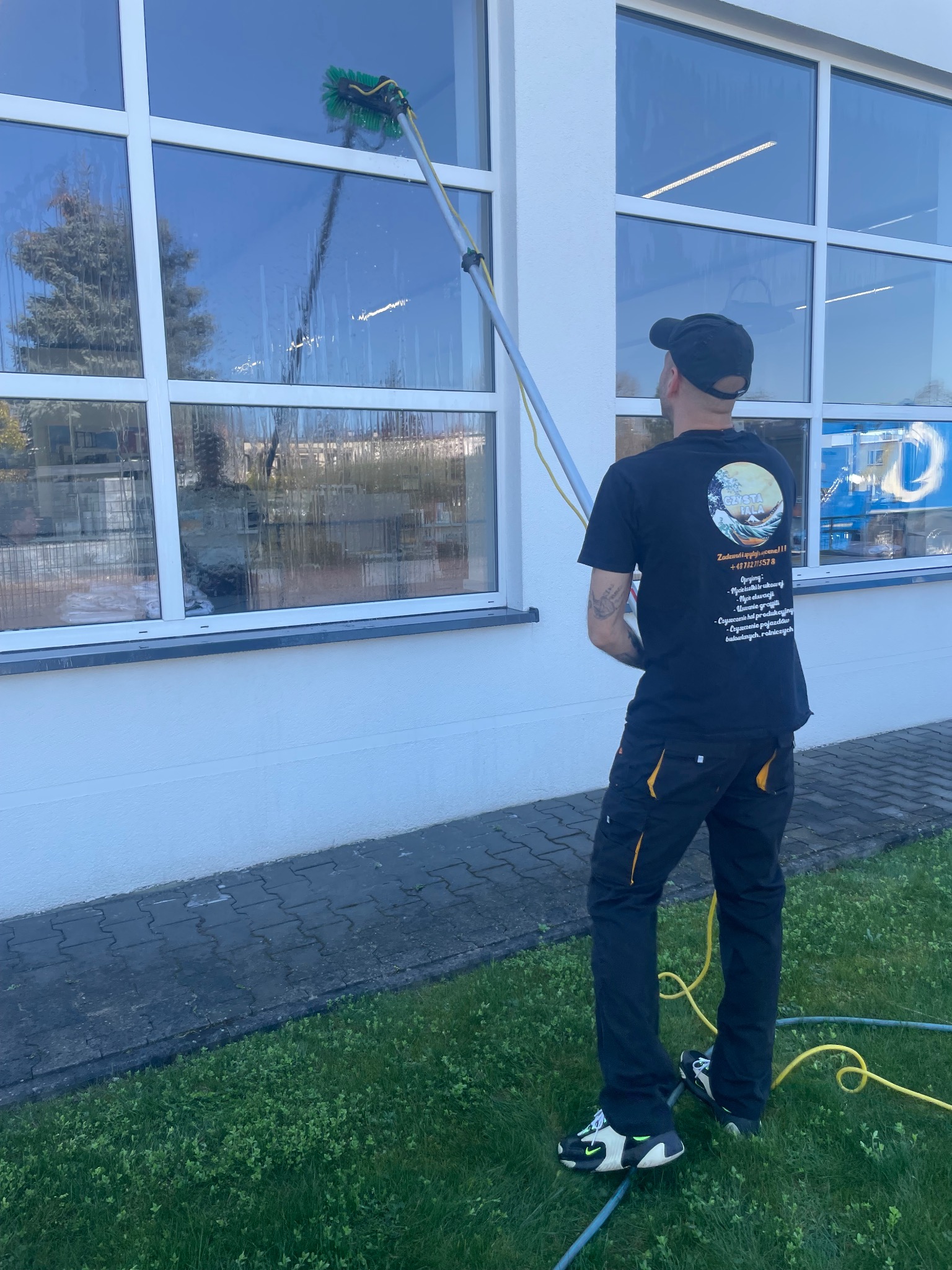 Pracownik w Poznaniu myje okna elewacji budynku za pomocą szczotki na teleskopowym kiju, widoczny fragment elewacji i trawnika, na plecach pracownika widoczne logo firmy 'Czysta Fala'.